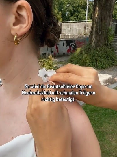 Braut wird das Hochzeitscape mit glitzernder Brosche Modell KANE an ihrem Kleid mit schmalen Traegern befestigt von MOANA
